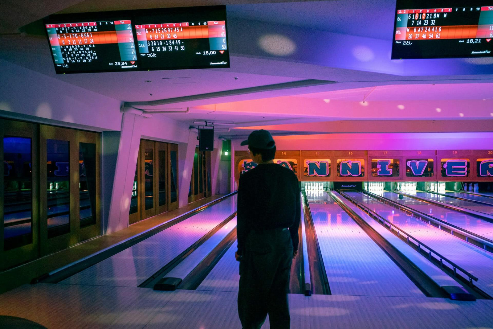 Semi-silhouetted individual standing in front of a bowling alley with dimmer lights for a dramatic effect. Above the individual and the lanes are screens showing bowling scores.