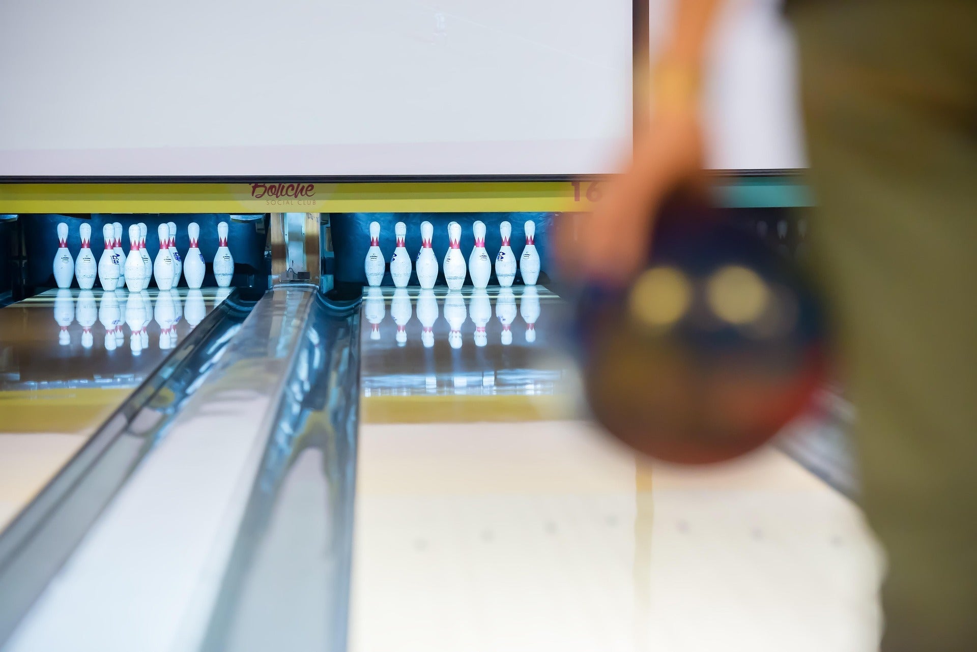 In the foreground, a left-handed person is holding a bowling ball in front of bowling pins